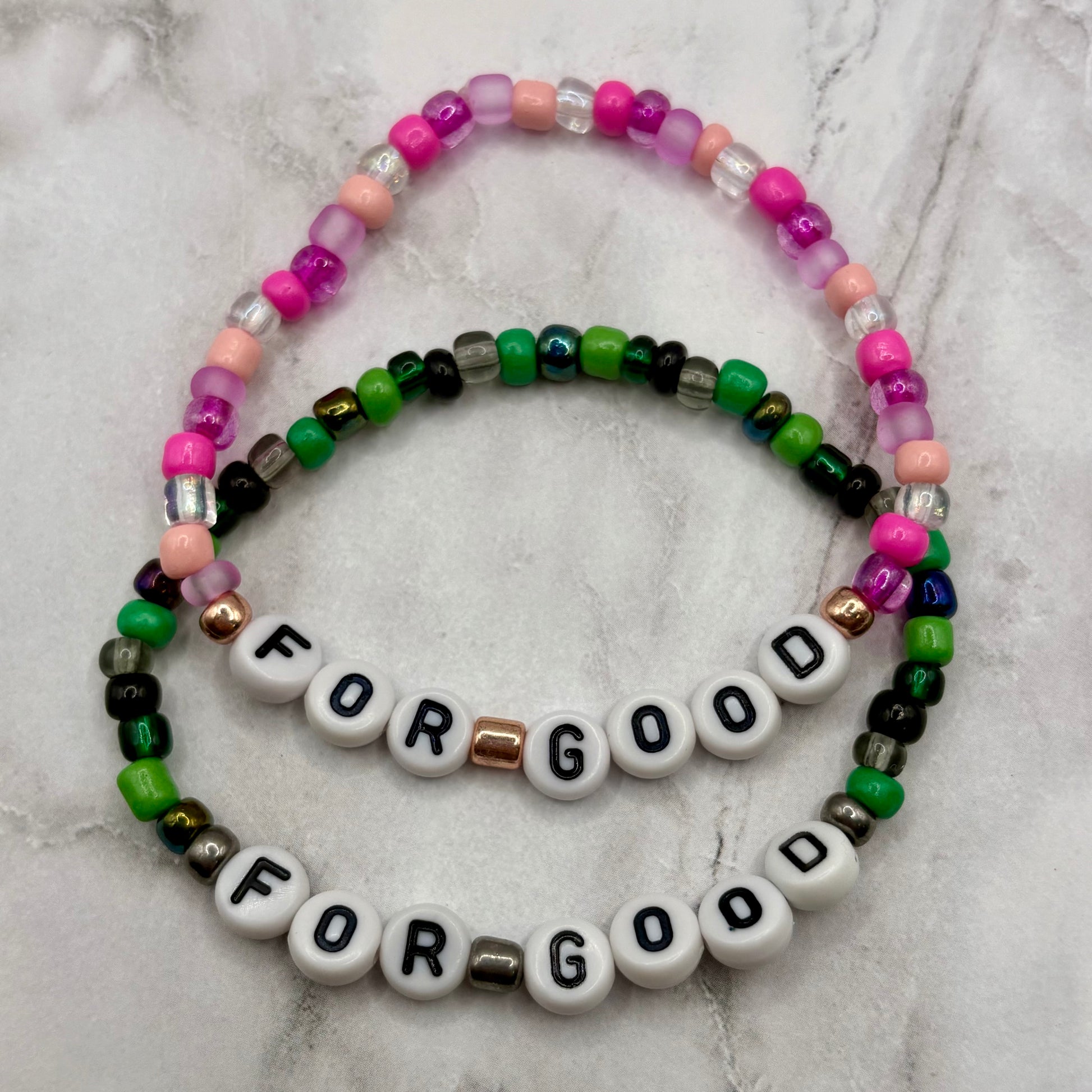 Two beaded bracelets with 'FOR GOOD' on a marble surface